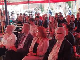 Pokaż zdjęcie: Na ławkach i krzesłach siedzą w rzędach ludzie, którzy oglądają występy na scenie. 