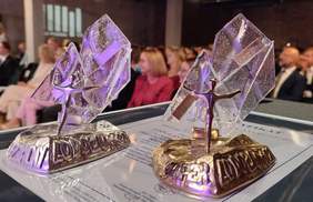 Pokaż zdjęcie: Na stoliku stoją dwie statuetki. W tle widać ludzi na widowni siedzących na krzesłach. 