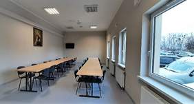 Pokaż zdjęcie: Jadalnia, do uzytku wspólnego, dwa długie, w kolorze jasnego drewna stoły, wokół poustawiane czaren krzesła (około 30), na scianie po lewej stronie wisi obraz z wizerunkiem świętego, po prawej stronie cztery duze okna i pod nimi kaloryfery, całość bardzo jasna i przestonna