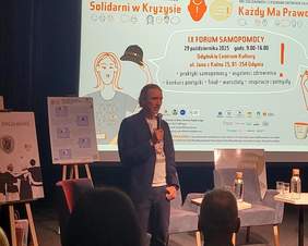 Pokaż zdjęcie: Mężczyzna z mikrofonem w dłoni, obok stoliki z wodą i krzesła; w tle ilustracja postaci oraz treść wydarzenia z datą 29 października 2025.