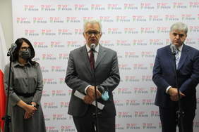 Pokaż zdjęcie: Na tle ścianki z logotypami PFRON stoją trzy osoby. Mężczyzna stojący w środku mówi do mikrofonu. Wszyscy są elegancko ubrani. Kobieta z lewej w garsonce, mężczyźni w garniturach.