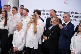 Pokaż zdjęcie: Grupa osób pozuje do zdjęcia. Osoby po lewej stronie są ubrane w białe koszule z godłem Polski na lewej piersi. Po prawej stronie stoją osoby ubrane w garnitury i eleganckie żakiety. W tle widać baner Igrzysk Paraolimpijskich w Mediolanie i Cortinie. 