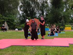 Pokaż zdjęcie: Na trawie rozłożona jest różowa mata na środku maty stoi dwóch aktorów ubranych na czarno twarze maja zasłonięte przyłbicami między nimi stoi chłopak ubrany w czarne spodnie i szarą bluzę z kapturem który jest założony na głowę na ramio