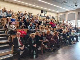 Pokaż zdjęcie: sala konferencyjna, na krzesłach siedzą osoby i  wpatrują się w scenę na której  prezentowane są prezentacje omawiane przez prelegentów konferencji