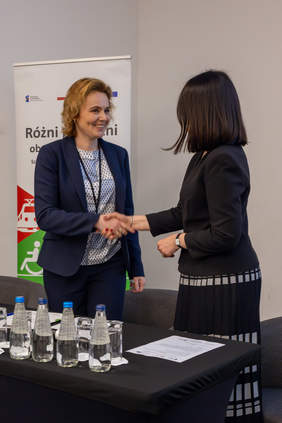 Pokaż zdjęcie: Dwie kobiety przedstawicielka Kolei Małopolskich p. Iwona Zwierzyk-Klimek i Wiceprezes PFRON p. Dorota Habich gratulują sobie, ściskając sobie dłonie. W tle baner z napisem Różni podróż – obsługa bez barier.