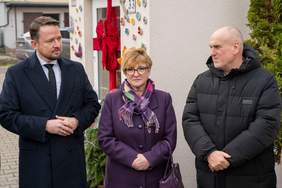 Pokaż zdjęcie: Trzy osoby stoją na zewnątrz. Po lewej stoi mężczyzna w ciemnym płaszczu i krawacie. W centrum stoi kobieta w fioletowym płaszczu i wielobarwnym szalu. Po prawej stoi mężczyzna w czarnej, puchowej kurtce z kapturem. Wszyscy trzej patrzą w prawą stronę. Za nimi widoczna jest ozdobiona ściana budynku.