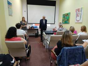 Pokaż zdjęcie: W sali siedzą na krzesłach różne osoby. Przed zebranymi stoi pan w garniturze. W tle widać rzutnik. 