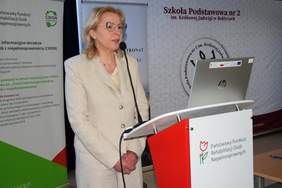 Pokaż zdjęcie: Przy mównicy stoi kobieta w beżowym garniturze.