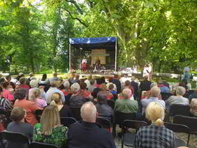Wśród drzew stoi scena, na której występują dwie kobiety. Przed sceną siedzą na krzesłach uczestnicy wydarzenia, widzimy ich z tyłu
