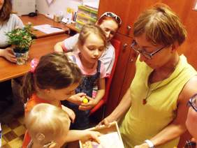 Pokaż zdjęcie: Trójka dzieci pokazujące pudełko osobie dorosłej. Przy biurku siedzi osoba dorosła trzymająca w rękach doniczkę z kwiatkiem