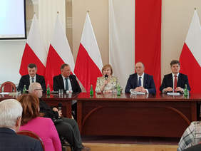 Grupa kilku osób, kobiet i mężczyzn przy stole prezydialnym, za nimi flagi biało-czerwone, widać także zebranych gości, którzy siedzą
