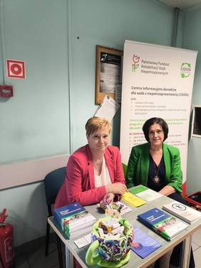 Pokaż zdjęcie: Dwie osoby siedzą przy stoliku informacyjnym PFRON i CIDON; na stole ulotki, broszury i materiały edukacyjne, w tle roll-up z informacjami o wsparciu dla osób z niepełnosprawnościami.
