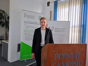 Pokaż zdjęcie: Kobieta blondynka, włosy upięte, w okularach, ubrana w ciemny garnitur stoi przy mównicy.