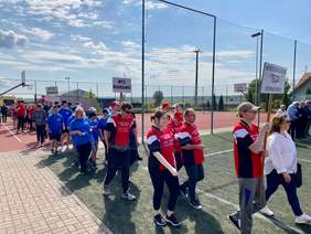 Pokaż zdjęcie: Zawodnicy z różnych placówek aktywizujących osoby z niepełnosprawnością mają na sobie koszulki z napisem danej placówki. Osoby te idą w jednym rzędzie przez dwa boiska. Każda drużyna niesie tabliczkę z nazwą placówki. W tle lekko zachmurzone niebo.