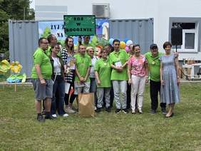 Pokaż zdjęcie: Grupa kilkunastu osób stoi na trawniku przed budynkiem i metalowym kontenerem, na którym wisi baner promujący imprezę sportową. Część osób trzyma w ręce nagrody lub dyplom. Z kolei jeden z mężczyzna trzyma tabliczkę z napisem: „ŚDS w Rogoźnie”.