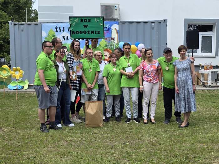 Pokaż zdjęcie: Grupa kilkunastu osób stoi na trawniku przed budynkiem i metalowym kontenerem, na którym wisi baner promujący imprezę sportową. Część osób trzyma w ręce nagrody lub dyplom. Z kolei jeden z mężczyzna trzyma tabliczkę z napisem: „ŚDS w Rogoźnie”.