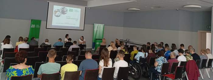 Pokaż zdjęcie: Grupa około czterdziestu osób siedzi w dużej sali i przysłuchuje się wystąpieniu dwóch kobiet. Z przodu, po obu stronach sali, ustawione są dwa banery ZUS, a na ekranie wyświetlony został slajd promujący Dni Osób z Niepełnosprawnościami.