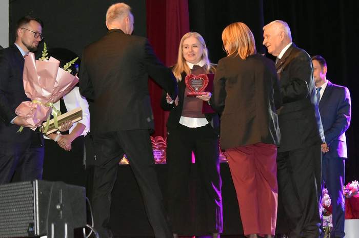 Pokaż zdjęcie: Grupa osób w formalnych strojach stoi na scenie. Kobieta w centrum, ubrana w ciemne spodnie i sweter na jasnej koszuli, uśmiecha się, trzymając statuetkę w kształcie serca. Wokół niej stoją mężczyźni i kobiety, prawdopodobnie gratulując jej. Jeden z mężczyzn po lewej trzyma duży bukiet różowych kwiatów.
