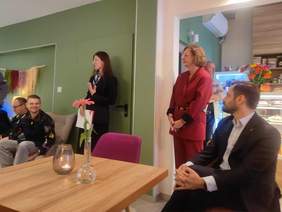 Pokaż zdjęcie: Przestrzeń Kawiarni, na środku stoją dwie kobiety, jedna przemawia. Po bokach przy stolikach siedzą mężczyźni, którzy słuchają. 