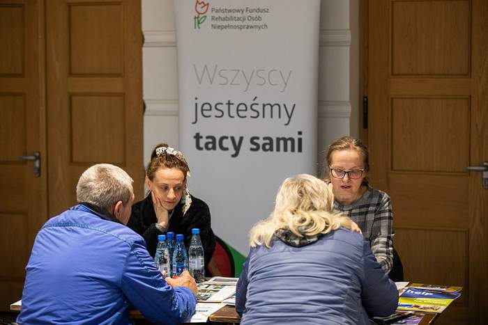 Pokaż zdjęcie: Przy stoliku siedzą trzy kobiety i mężczyzna w tle widać dwoje dużych drzwi a po środku nich baner z logo PFRON i napisem wszyscy jesteśmy tacy sami
