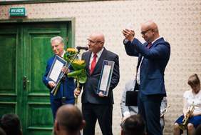 Pokaż zdjęcie: Dwóch łysych panów w garniturach i krawatach, jeden z uniesionymi rękoma w geście zwycięstwa i radości