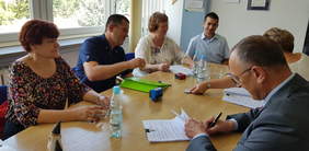 6 osób siądzacych przy jasnym stole, 3 kobiety, 3 mężczyzn, w trakcie podpisania umowy, osoby usmiechają się, na stole leżą dokumenty, pieczątki, stoi woda w butelkach i szklankach