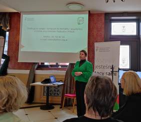 Pokaż zdjęcie: Kobieta w zielonym sweterku przemawia do zgromadzonych osób za nią na ekranie wyświetla się prezentacja.