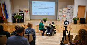 Pokaż zdjęcie: Dwie kobiety, jedna z nich siedzi na wózku inwalidzkim przemawia do osób siedzących naprzeciwko, druga z kobiet w dłoni trzyma mikrofon skierowany do przemawiającej, za nimi na ekranie wyświetla się napis Konferencja O oswajaniu inkluzji w rzeczywi