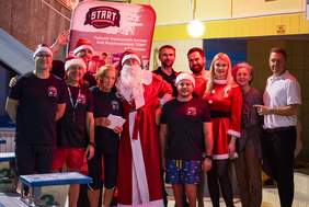 Pokaż zdjęcie: Grupa osób, w tym trenerzy i organizatorzy zawodów pływackich, pozuje do zdjęcia w świątecznych strojach na tle baneru Podlaskiego Stowarzyszenia Sportowego Osób Niepełnosprawnych "START" w Białymstoku. W centrum znajduje się osoba przebrana za Świętego Mikołaja oraz kobieta w sukience Mikołajkowej.