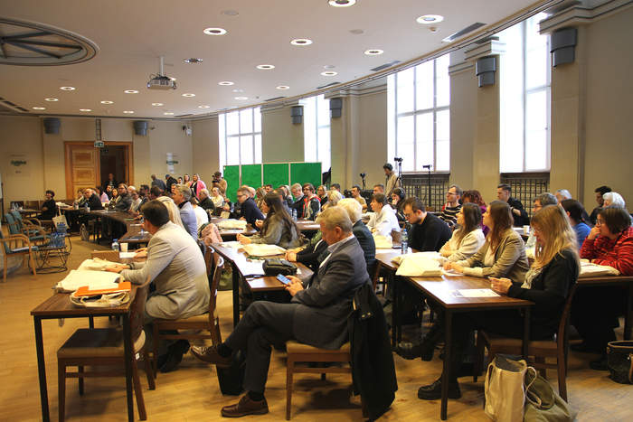 Pokaż zdjęcie: Na zdjęciu widoczna sala konferencyjna wypełniona ludźmi siedzącymi przy stołach. Duża jasna sala z wysokimi oknami po prawej stronie i sufitowymi punktowymi lampami. 