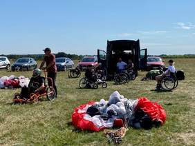 Pokaż zdjęcie: Na zielonej trawie  leży paralotnia i opiekun prowadzi wózek. Trwają przygotowania do szkolenia z latania na paralotni. Opiekunowie przygotowują sprzęt do latania.