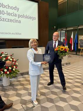 Pokaż zdjęcie: Barbara Jaskierska podczas odbierania tytułu Honorowego Ambasadora Szczecina. Ubrana w jasny garnitur, trzyma w rękach statuetkę i dyplom. Obok niej Prezydent Szczecina wręcza bukiet kwiatów. W tle ekran z napisem "Obchody 80-lecia polskiego Szczecina".