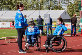 Pokaż zdjęcie: Na bieżni dwie zawodniczki siedzą na wózkach inwalidzkich. Jedna  nich naciąga gumę do ćwiczeń z inna zawodniczką w ramach ćwiczeń.