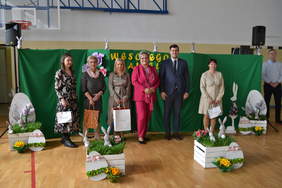 Pokaż zdjęcie: Na zdjęciu znajduje się pięć kobiet i mężczyzna. Cztery kobiety trzymają w rękach torby z upominkami. Stoją na tle zielonego parawanu