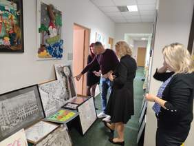 Pokaż zdjęcie: W długim korytarzu, po jego lewej i prawej stronie na podłodze i na krzesłach poustawiane są prace plastyczne. Pośród prac spaceruje komisja, która ocenia prace w poszczególnych kategoriach. Na zdjęciu widać cztery osoby.