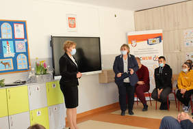 Pokaż zdjęcie: Dwie kobiety stoją na tle czarnej tablicy, przemawiają do siedzących uczestników spotkania