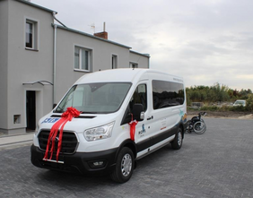 Pokaż zdjęcie: biały, 9-cioosobowy bus, z napisem MOPS Żnin i udekorowany czerwonymi wstęgami, na tle szarego budynku