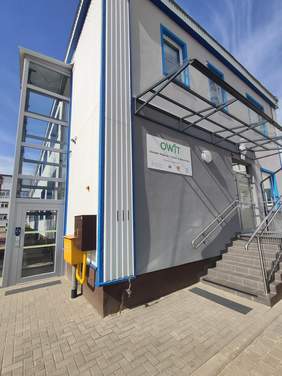 Pokaż zdjęcie: Nowoczesny, dwukondygnacyjny budynek z szarymi ścianami i niebieskimi akcentami wokół okien i krawędzi. Na ścianie widać szyld z napisem "OWiT". Przy budynku znajdują się schody prowadzące do wejścia głównego, z poręczami po obu stronach. Z boku widoczna jest winda, zapewniająca dostępność dla osób z niepełnosprawnościami. Całość jest otoczona kostką brukową.