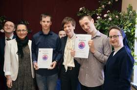 Zdjęcie grupowe sześciu osób stojących obok siebie na tle jasnej ściany. W centrum dwaj mężczyźni prezentują dyplomy z napisem „Dyplom” i kolorową grafiką. Od lewej: kobieta w okularach i jasnej marynarce, młody mężczyzna w granatowej koszuli, w tle twarz kolejnej osoby, starsza kobieta w białej koszuli i ciemnej marynarce, młody mężczyzna w szarej koszuli oraz zakonnica w granatowym habicie. Wszyscy są uśmiechnięci.