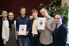 Pokaż zdjęcie: Zdjęcie grupowe sześciu osób stojących obok siebie na tle jasnej ściany. W centrum dwaj mężczyźni prezentują dyplomy z napisem „Dyplom” i kolorową grafiką. Od lewej: kobieta w okularach i jasnej marynarce, młody mężczyzna w granatowej koszuli, w tle twarz kolejnej osoby, starsza kobieta w białej koszuli i ciemnej marynarce, młody mężczyzna w szarej koszuli oraz zakonnica w granatowym habicie. Wszyscy są uśmiechnięci.