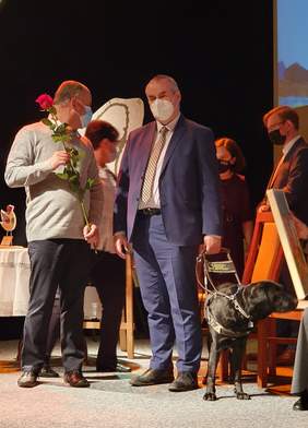 Pokaż zdjęcie: Paweł Wdówik, wiceminister, pełnomocnik rządu ds. osób niepełnosprawnych. Pełnomocnik stoi na scenie, towarzyszy mu pies przewodnik.