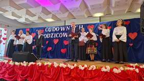 Pokaż zdjęcie: Grupa osób stoi na scenie śpiewa do mikrofonów. W tle widoczny jest granatowy baner z napisem „Cudownych Rodziców Mam” oraz dekoracje z czerwonych serc.