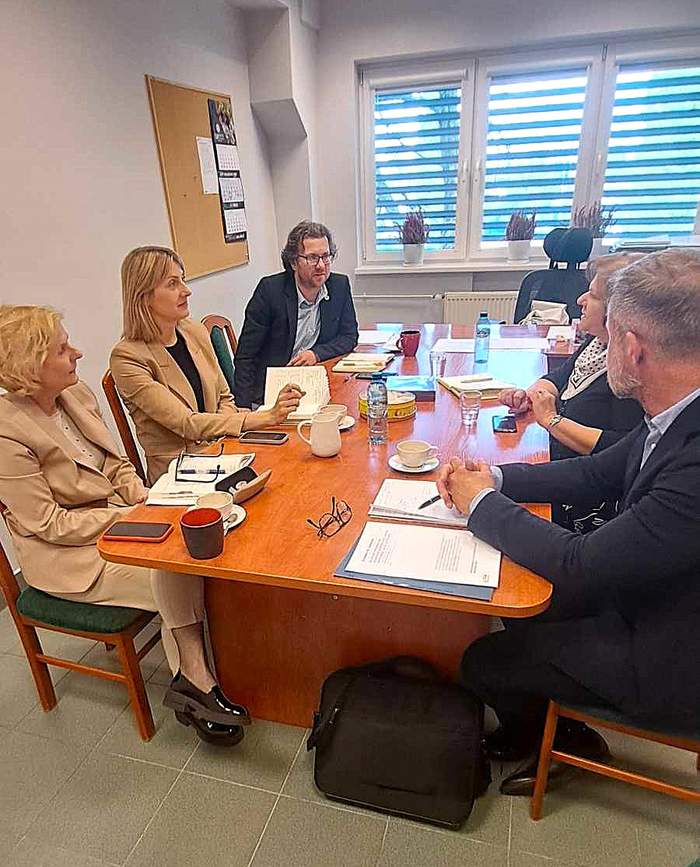 W gabinecie, wokół stołu konferencyjnego siedzi 5 osób - trzy kobiety i dwóch mężzyzn i rozmawiają. Na stole leżą otwarte notesy z długopisami, telefony i okulary oraz stoją filiżanki i woda w butelkach.