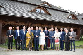 Pokaż zdjęcie: Uczestnicy Konwentu Powiatów Województwa Małopolskiego. Uśmiechnięta, liczna grupa osób pozuje do zdjęcia. W tle drewniany budynek.