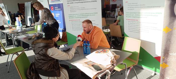 Przy stoliku, w punkcie informacyjnym PFRON siedzi mężczyzna w pomarańczowym swetrze i udziela wywaidu redaktorce z Radia PiK, która siedzi naprzeciwko niego i trzyma w dłoni mikrofon z logo radia