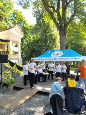 Pokaż zdjęcie: Na scenie trwa występ jednej z grup, większość aktorów ubrana jest na biało czarno, ustawieni są w półkole.