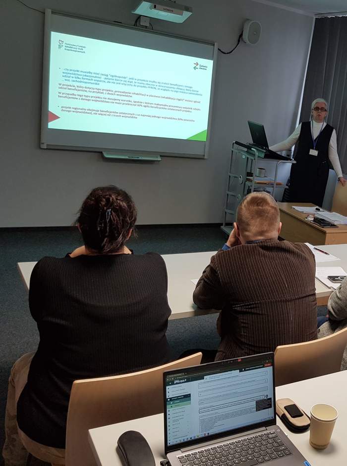 Pokaż zdjęcie: Grupa kilku osób siedzi za ustawionymi w dwóch rzędach stolikami. Część z tych osób posiada laptop ustawiony na stoliku, natomiast pozostała grupa ma przed sobą notatki w formie papierowej. Z przodu sali widoczna jest jedna kobieta prowadząca szkolenie oraz ekran, na którym wyświetlana jest prezentacja.