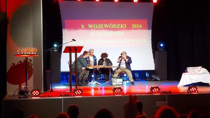 Pokaż zdjęcie: Na scenie siedzi przy stoliku grupa czterech mężczyzn. Jeden z mężczyzn gestykuluje rękoma. W tle, na dużym ekranie wyświetlony jest napis „X Wojewódzki Kabareton Osób Niepełnosprawnych 2024”. Po lewej stronie, na okrągłym stoliku ustawione są statuetki.
