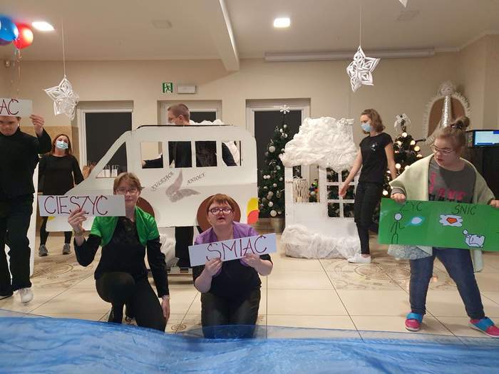 Pokaż zdjęcie: W dużym pomieszczeniu 2 dziewczyny klęczą trzymają w rękach białe kartki z napisem cieszyć się, śmiać obok stoi dziewczyna trzyma zielona kartkę z napisem żyć śnić. Na drugim planie chłopak trzyma w rękach duży papierowy szablon busa, ob