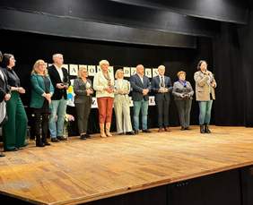 Pokaż zdjęcie: Widzimy na drewniane scenie 11 stojących osób, wypowiada się Zastępca Dyrektora Oddziału PFRON w Szczecinie. W tle czarny napis na białym tle "Światowy Dzień Warsztatów Terapii Zajęciowej"